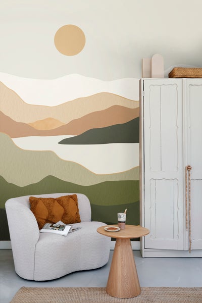 Helle, freundliche Inneneinrichtung mit Sessel, Tisch und Wandschrank vor einer Wandtapete mit Landschaftsmotiv.