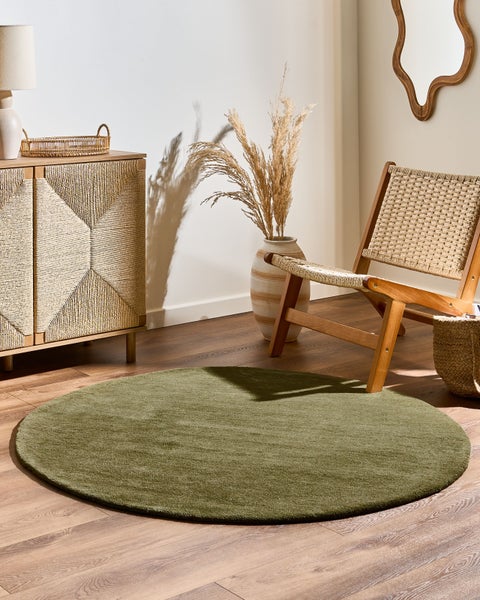 Runder grüner Teppich auf Holzboden in hellem Raum mit Holzkommode, Lounge-Sessel und Vase mit Trockenblumen im natürlichen Stil.