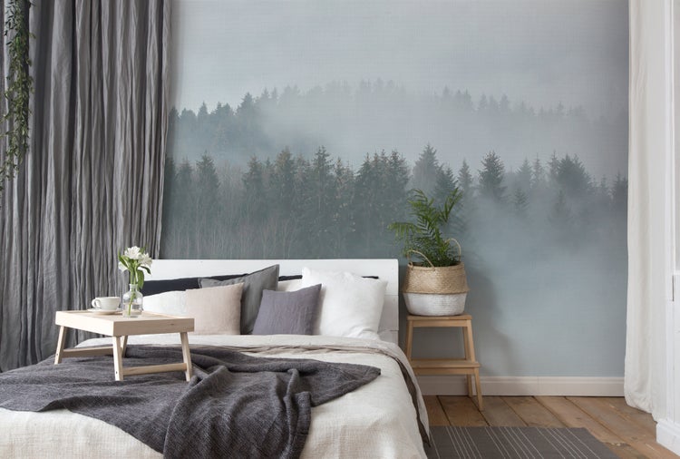 Schlafzimmer mit Fototapete eines nebligen Waldes, Bett mit Kissen, Betttablett aus Holz und Zimmerpflanze auf einem Schemel.