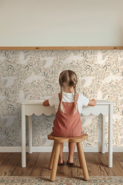 Mädchen sitzt an einem weißen Tisch vor einer Tapete mit floralem Muster und Schwanmotiven