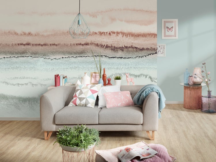 Modernes Wohnzimmer mit einer Vliestapete im abstrakten Aquarell-Design, einem grauen Sofa und hellem Boden in Holzoptik.
