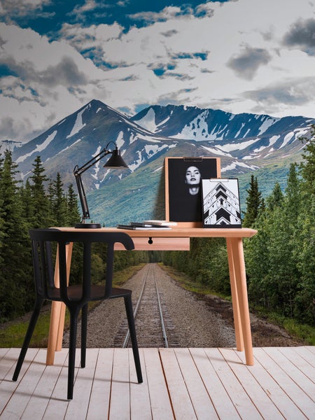 Fototapete mit Gebirgslandschaft und Eisenbahngleisen hinter einem Holzschreibtisch mit modernem Stuhl und einer Schreibtischlampe.