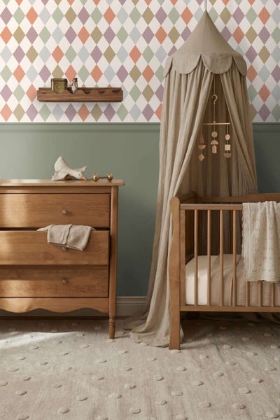 Einrichtung eines Kinderzimmers mit Holzkommode, Gitterbett mit Betthimmel, geometrischer Tapete in Rautenmuster und einem getufteten Teppich.