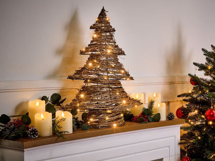 Dekorativer beleuchteter Weihnachtsbaum aus Zweigen mit Kerzen auf Kaminsims neben geschmücktem Weihnachtsbaum.