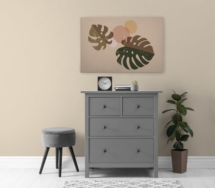 Eine Kommode mit drei Schubladen neben einem Hocker und einer Zimmerpflanze unter einem Wandbild mit abstrakten Pflanzenmotiven.