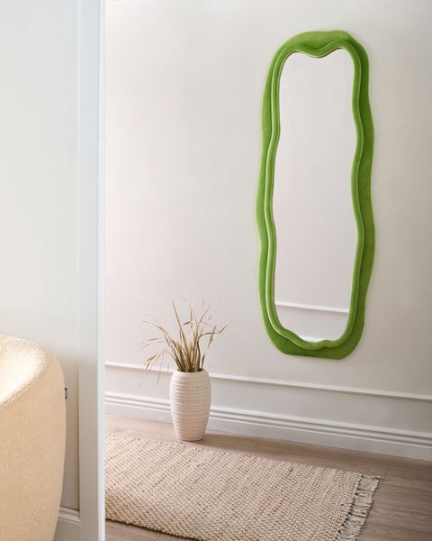 Raumansicht mit einem unregelmäßig geformten Spiegel mit grünem Rahmen, einer Vase mit getrockneten Pflanzen und einem Teppich