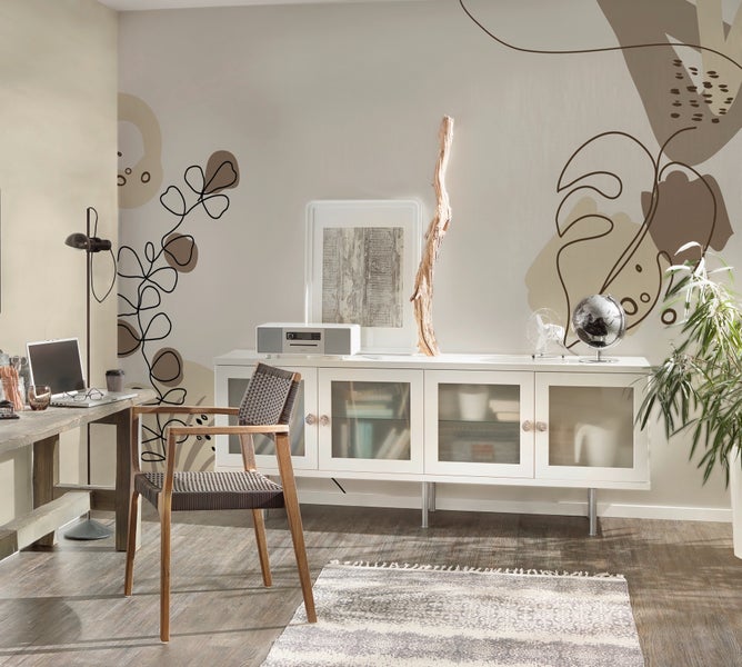 Modernes helles Zimmer mit abstrakter botanischer Wandtapete, weißem Sideboard, geflochtenem Holzstuhl und hölzernem Schreibtisch mit Laptop.