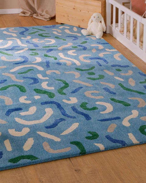 Blauer Kinderteppich mit abstraktem Muster auf einem Holzboden in einem hellen Kinderzimmer neben einem weißen Gitterbett.