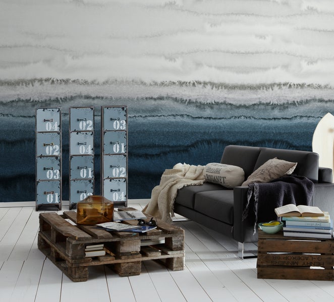 Industrielles Wohnzimmer mit dunklem Sofa, Couchtisch aus Paletten, Metall-Kommoden und Tapete im Aquarell-Stil auf hellem Holzfußboden.