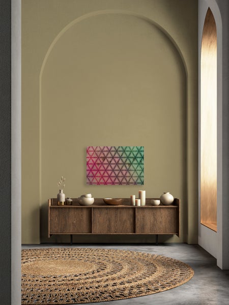 Wohnzimmer mit dunkler Holzkommode, rundem Jute Teppich, Kerzen und Vasen vor einer Wand mit dekorativem Wandbild in Dreiecksoptik.