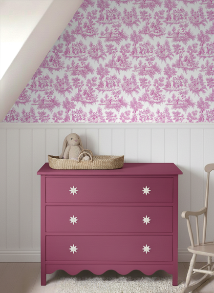 Kinderzimmer mit rosa Kommode vor einer Wand mit weißen Paneelen und rosa Toile-de-Jouy-Tapete. Auf der Kommode steht ein Weidenkorb.