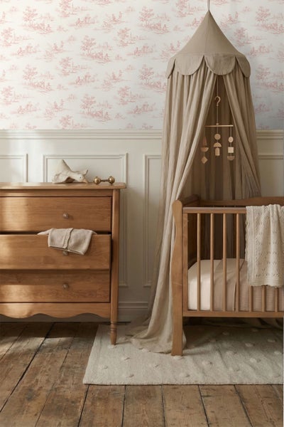 Babyzimmer mit Vliestapete im Toile-de-Jouy-Stil, Holzkommode, Kinderbett mit Betthimmel und Wandtäfelung auf Holzdielen.