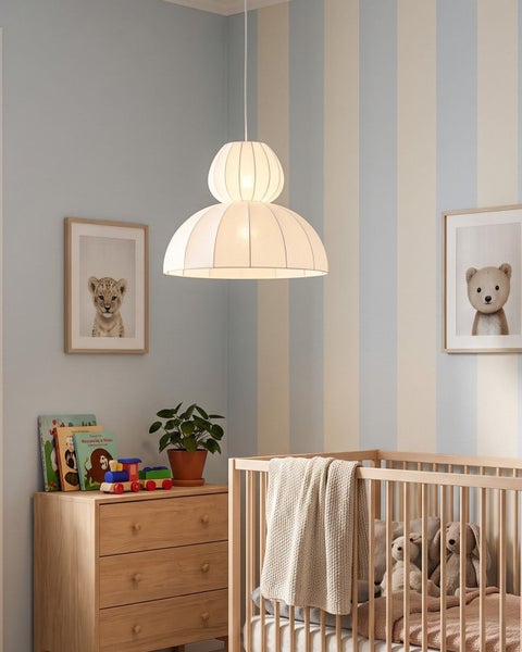 Ein Kinderzimmer mit einer modernen runden Textilpendelleuchte, einem Holzkinderbett, einer Kommode, Wandbildern und gestreiften Tapeten.