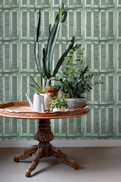 Dekorative Zimmeransicht mit Pflanzen auf einem Tisch vor einer Wandtapete mit Fensterladenmotiv.