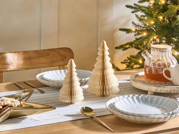 Festlich gedeckter Tisch mit Papierweihnachtsbäumen, Geschirr und Weihnachtsbaum im Hintergrund.