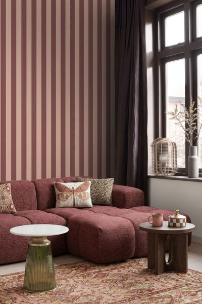 Wohnzimmer mit rot-rosa gestreifter Tapete, rotem Modulsofa, Zierkissen, runden Beistelltischen und Fenster mit dunklen Vorhängen.