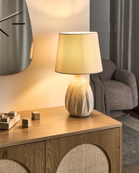 Hell beleuchtete Szene mit einer Tischleuchte aus Keramik und Stoff auf einem Sideboard aus Holz und Rattan neben einem Sessel.