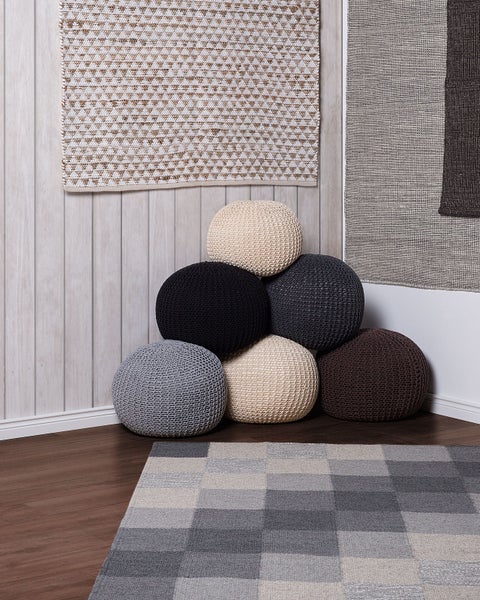 Mehrere gestrickte Poufs und ein karierter Teppich in einem hellen Raum mit Holzpaneelen und dunklem Holzboden.