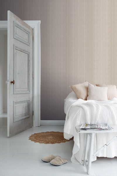 Helles Schlafzimmer mit dezent gemusterter Tapete, weißem Bett mit Strickdecke, kleiner Holzbank und rustikaler Zimmertür.