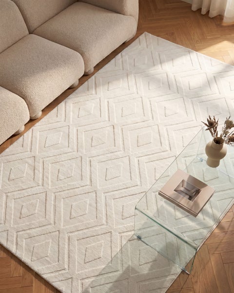 Wohnzimmer mit beigefarbenem Sofa, einem rechteckigen Teppich mit geometrischem Rautenmuster und einem Couchtisch aus Glas auf Parkettboden.