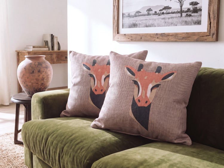 Zwei Dekokissen mit Giraffenmotiv auf einem Sofa in einem Wohnzimmer mit einer Vase und einem gerahmten Bild einer Savanne.