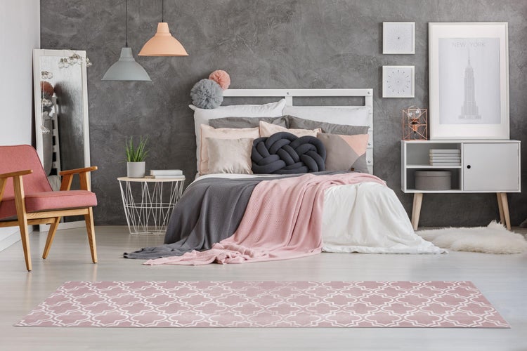 Modernes helles Schlafzimmer mit grauer Strukturwand, weißem Bett, rosa und grauen Textilien, Teppich, Sessel und dekorativer Beleuchtung.