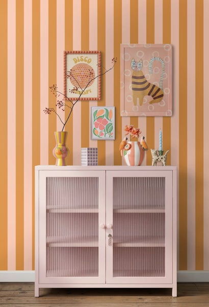 Vliestapete mit vertikalen Streifen in Orange und Rosa hinter einem rosa Schrank mit geriffelten Glastüren.