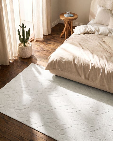 Helles Schlafzimmer mit weißem Teppich mit Blattmuster auf dunklem Holzboden, neben einem Bett und einem Kaktus am Fenster.