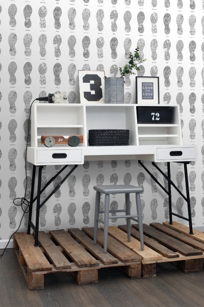 Weißer Schreibtisch mit schwarzem Gestell, grauer Hocker und dekorativen Elementen auf einer Holzpalette vor einer Tapete mit Fußabdruckmuster.