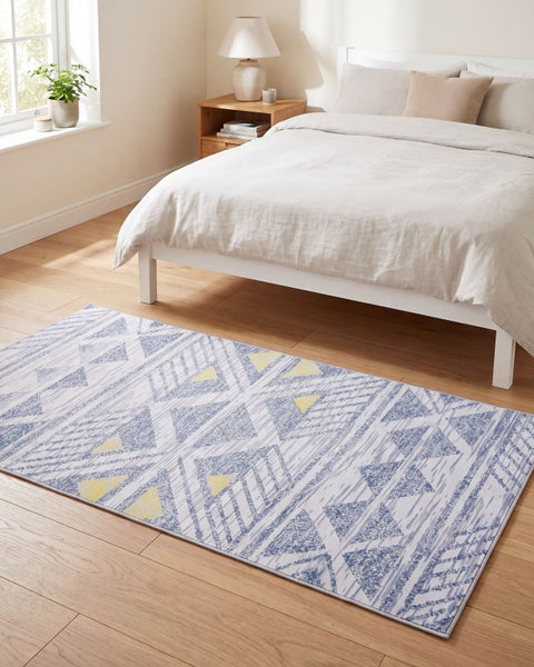 Ein rechteckiger Teppich mit geometrischem Muster liegt vor einem Bett in einem hellen Schlafzimmer mit Holzboden.