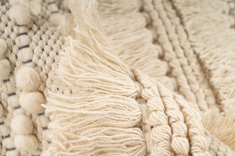 Nahaufnahme eines cremefarbenen Teppichs mit gewebter Struktur, Pompons und Fransen aus Wolle.