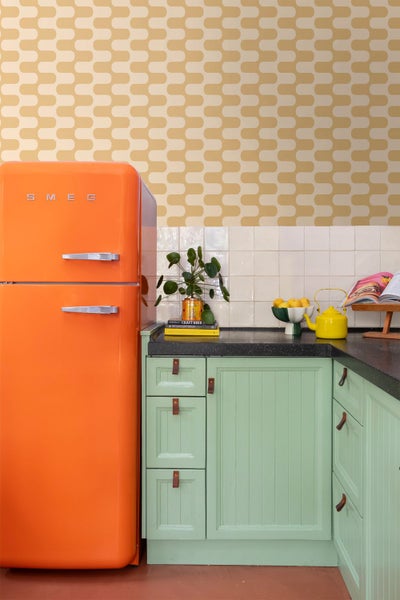 Helle Küche im Retro-Stil mit Smeg Logo auf orangefarbenem Kühlschrank, grünen Schränken mit Ledergriffen und Tapete mit Wellenmuster.