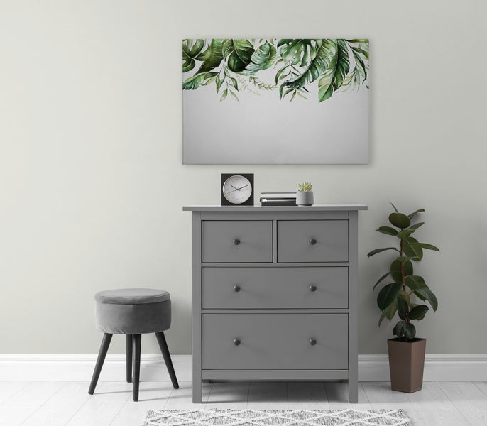 Wandbild mit tropischem Blattmotiv über einer grauen Kommode mit Hocker, Uhr und Pflanze in einem modern eingerichteten Wohnbereich.