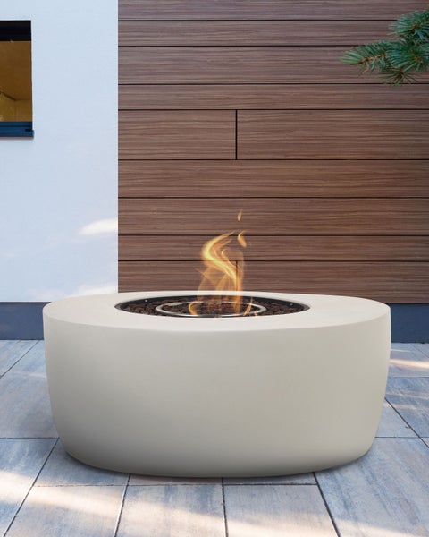 Moderne runde Gas-Feuerstelle aus hellem Faserbeton mit Flammen und Lavasteinen auf einer Terrasse vor einer Wand in Holzoptik.