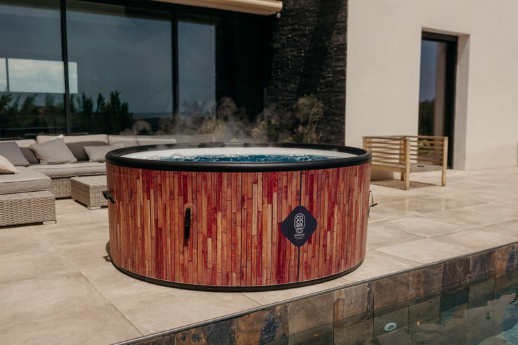 Aufblasbarer Whirlpool in Holzoptik auf einer Steinterrasse neben einem Pool, mit Gartenmöbeln im Hintergrund.