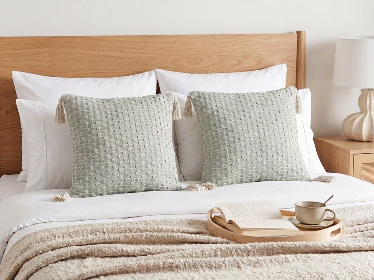 Hell eingerichtetes Schlafzimmer mit Holzbett, weißer Bettwäsche, zwei strukturierten grünen Zierkissen mit Quasten, beiger Kuscheldecke und Holztablett.