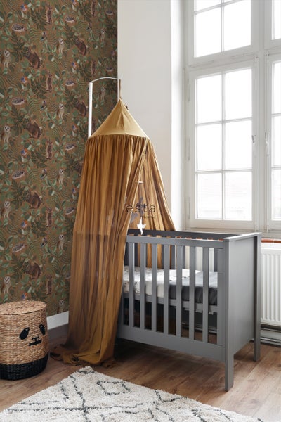 Ein Kinderzimmer mit einem grauen Kinderbett unter einem Baldachin, einem gemusterten Teppich und einer Korbtruhe vor einer Tapete mit Tiermotiven.