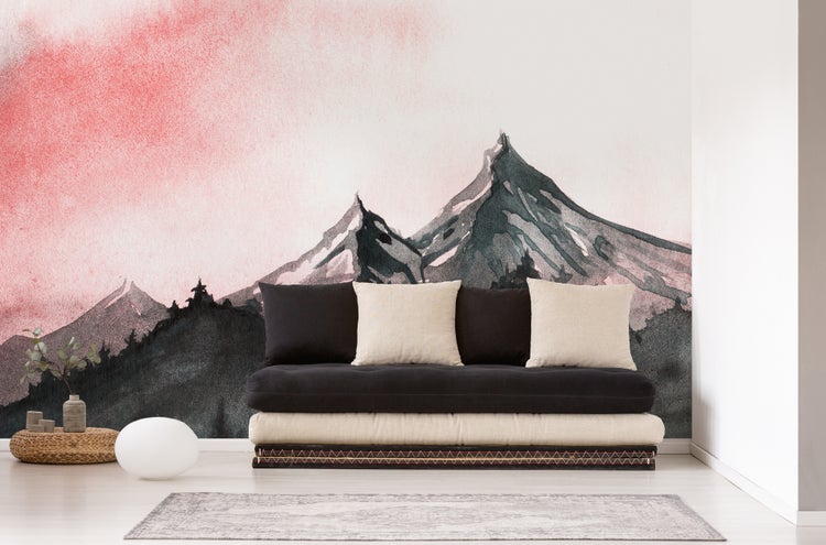 Fototapete mit Aquarell-Berglandschaft und rotem Himmel hinter einem modernen Futon-Sofa mit Kissen in einem hellen Wohnraum.