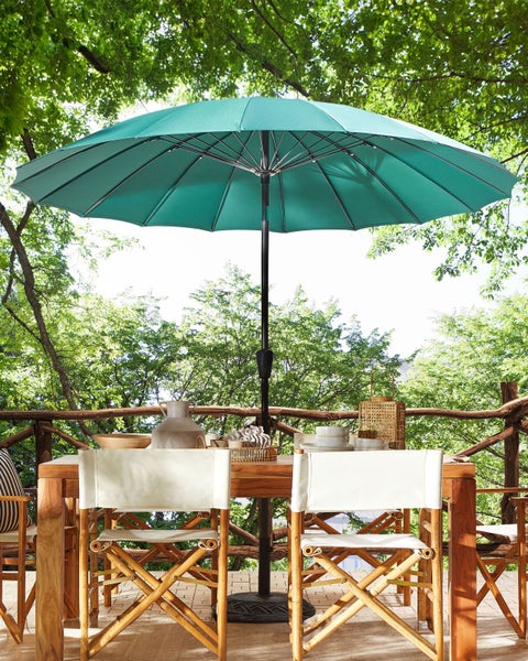 Großer türkisfarbener Sonnenschirm über einem hölzernen Gartentisch mit weißen Klappstühlen auf einer von Bäumen umgebenen Terrasse.