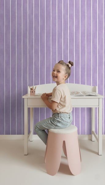 Mädchen sitzt an weißem Schreibtisch auf einem Hocker vor einer gestreiften Tapete