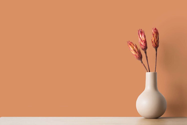 Beige Keramikvase mit drei getrockneten Blumen auf einem Holztisch vor einer apricotfarbenen Wand.
