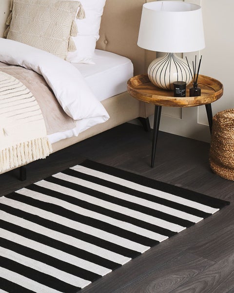 Schwarz-weiß gestreifter Teppich in einem organisierten Schlafzimmer neben einem Bett und runden Beistelltisch aus Holz auf dunklem Boden.