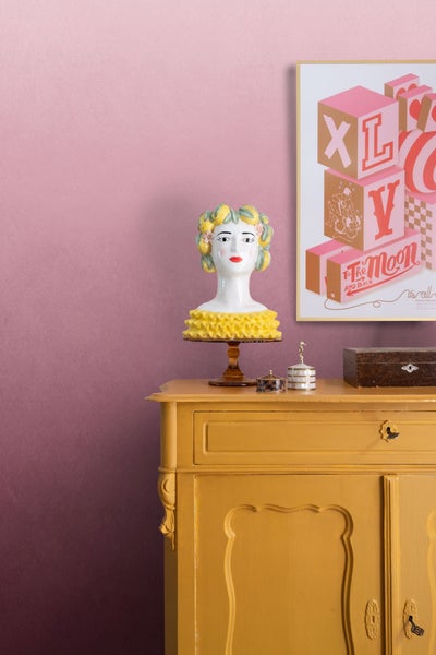 Dekorative Szene mit einer Frauenkopf-Skulptur auf einer Kommode neben einer gerahmten Illustration.