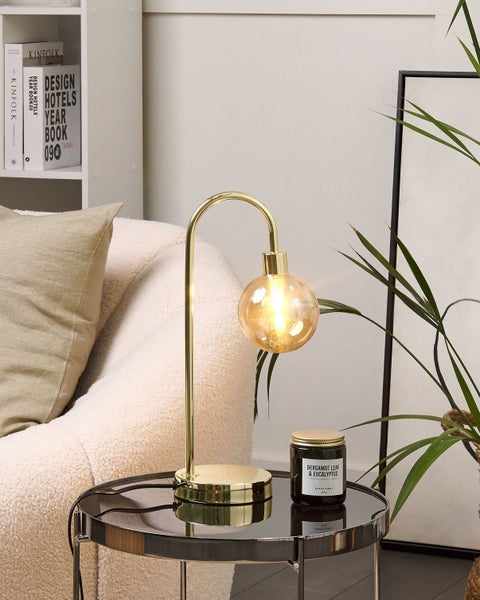 Dekorative Szene mit einer goldenen Lampe, einer Duftkerze und dekorativen Büchern auf einem Beistelltisch neben einem Sessel.