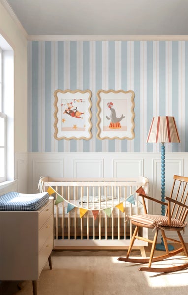 Hell eingerichtetes Kinderzimmer mit blau-creme gestreifter Tapete, weißem Gitterbett, hölzernem Schaukelstuhl und Bildern mit Zirkusmotiven.