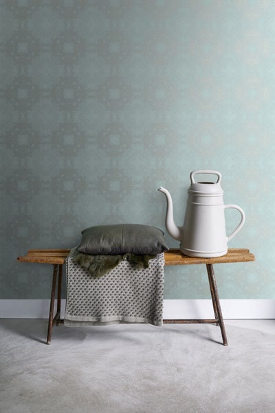 Dekoratives Stillleben mit Gießkanne, Kissen und Decke auf einer Holzbank vor einer verzierten Tapete.