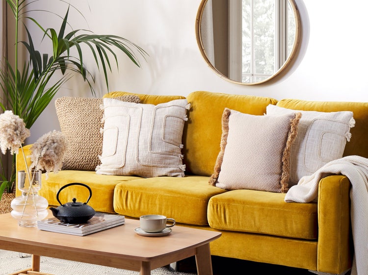 Wohnzimmer mit gelbem Sofa, Kissen, Holztisch, Teekanne und runden Spiegel.