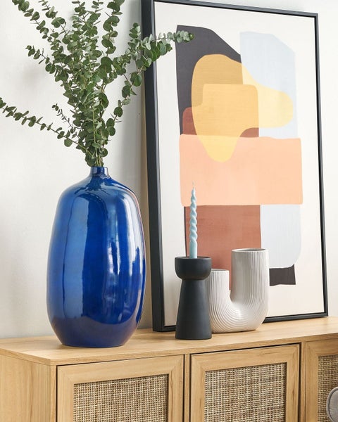 Dekorative Szene mit einer blauen Vase, Pflanzen, Kerzen und abstrakter Kunst auf einem Holzschrank.