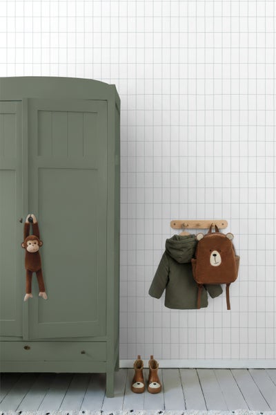 Hell gestaltetes Kinderzimmer mit weißer Gittertapete, einem grünen Kleiderschrank aus Holz sowie einer Garderobe mit Kinderjacke und Bärenrucksack.