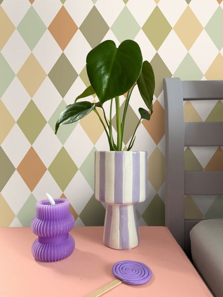 Dekorative Szene mit Zimmerpflanze in gestreiftem Übertopf, Lilafarbene Kerze und Kunst-Lutscher auf einem Tisch.